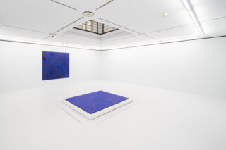 Ein heller Galerieraum mit weißem Boden und Wänden. An der Wand hängt ein blaues quadratisches Bild, am Boden liegt ein blaues quadratisches Bild.