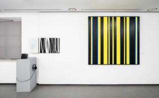 Bright gallery space featuring a large abstract artwork of black, yellow, and blue vertical stripes on the wall. A white pedestal with a tablet and headphones stands on the left.