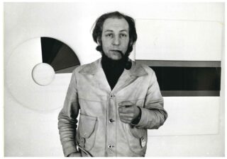 A young man with a pipe in his mouth, wearing a jacket, standing in front of his artworks.
