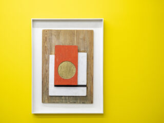 Abstract composition featuring a red rectangle with a gold circle, white and black rectangles, on a wood-grain background, in a white frame.