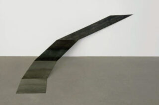 Black-and-white abstract sculpture with slanted, stepped structure against light gray wall and dark floor.