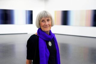 An older woman with medium-length gray hair, wearing a black top and a blue scarf, is standing in a modern gallery with abstract pictures on the walls.
