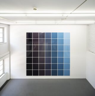 Large wall artwork composed of square panels in various shades of blue in a white room with tall windows.