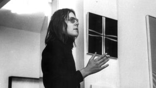 Black-and-white photo of a man with glasses and shoulder-length hair, standing sideways by a wall with abstract paintings, and gesturing with one hand.
