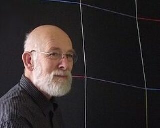 An older man with a gray beard and glasses standing in front of a black artwork.