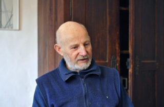 Older man with bald head and white beard wearing a dark blue fleece sweater sitting in front of a dark wooden cabinet.