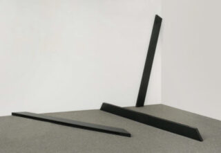 Three black bars of different lengths, lying and leaning on a gray floor agaist a white wall.