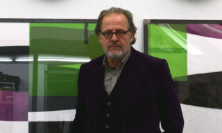 Man with glasses and beard standing in front of abstract, colorful paintings.