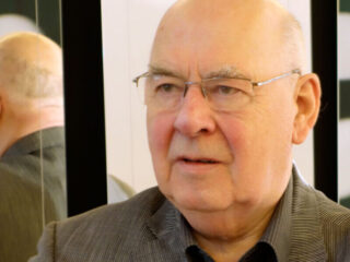 Older man wearing glasses and a gray blazer in front of a mirrored wall.