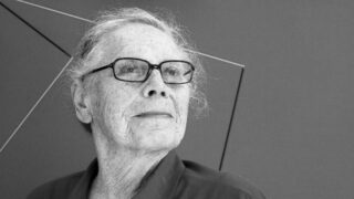An older woman with her gray hair pulled back, standing in front of an abstract artwork.