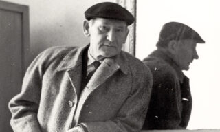 Older man wearing a flat cap and coat leaning against a marble fireplace with his reflection visible in the mirror.
