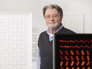 Älterer Mann mit Brille, grauem Haar und Bart, trägt Hemd und Pullover, steht vor zwei abstrakten Kunstwerken mit geometrischen Mustern.