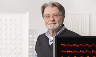 Older man with glasses, gray hair, and beard wearing shirt and sweater standing in front of two abstract artworks with geometric patterns.