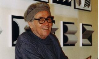Older man wearing glasses and a headband sitting with black-and-white geometric wall art behind.